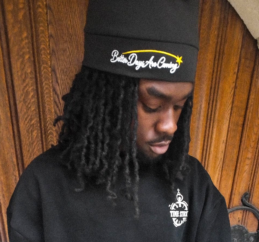 Person wearing a black sweatshirt with a logo and a black cap, standing in front of a wooden door.